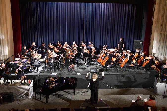 Bild eines Orchesters auf einer Bühne mit Dirigentin vor blauem Vorhang