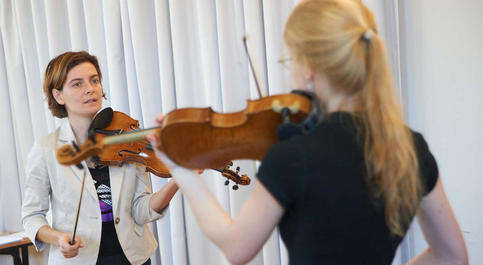 Abbildung: Teaserbild. Geigenlehrerin erklärt Studentin etwas am Instrument