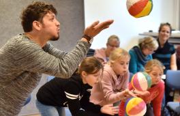 Junger Mann pustet gegen aufblasbaren Ball, eine Gruppe Kinder im Hintergrund ebenso