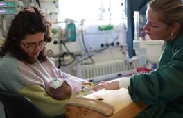 Musiktherapie in der Neonatologie; Musiktherapeutin mit Saiteninstrument am Bett einer Frau mit einem Neugeborenen