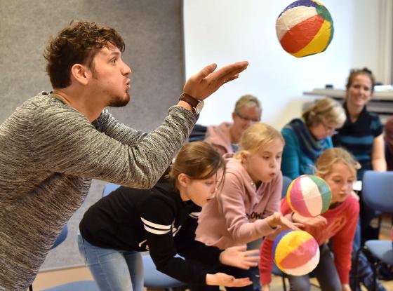 Junger Mann pustet gegen aufblasbaren Ball, eine Gruppe Kinder im Hintergrund ebenso