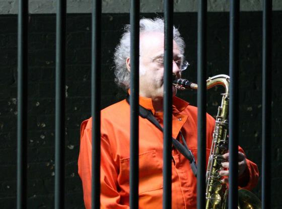 Älterer Herr in orangefarbenem Häftlingsoverall spielt Saxofon hinter Gittern