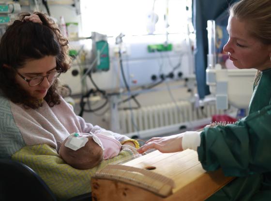 Musiktherapie in der Neonatologie; Musiktherapeutin mit Saiteninstrument am Bett einer Frau mit einem Neugeborenen